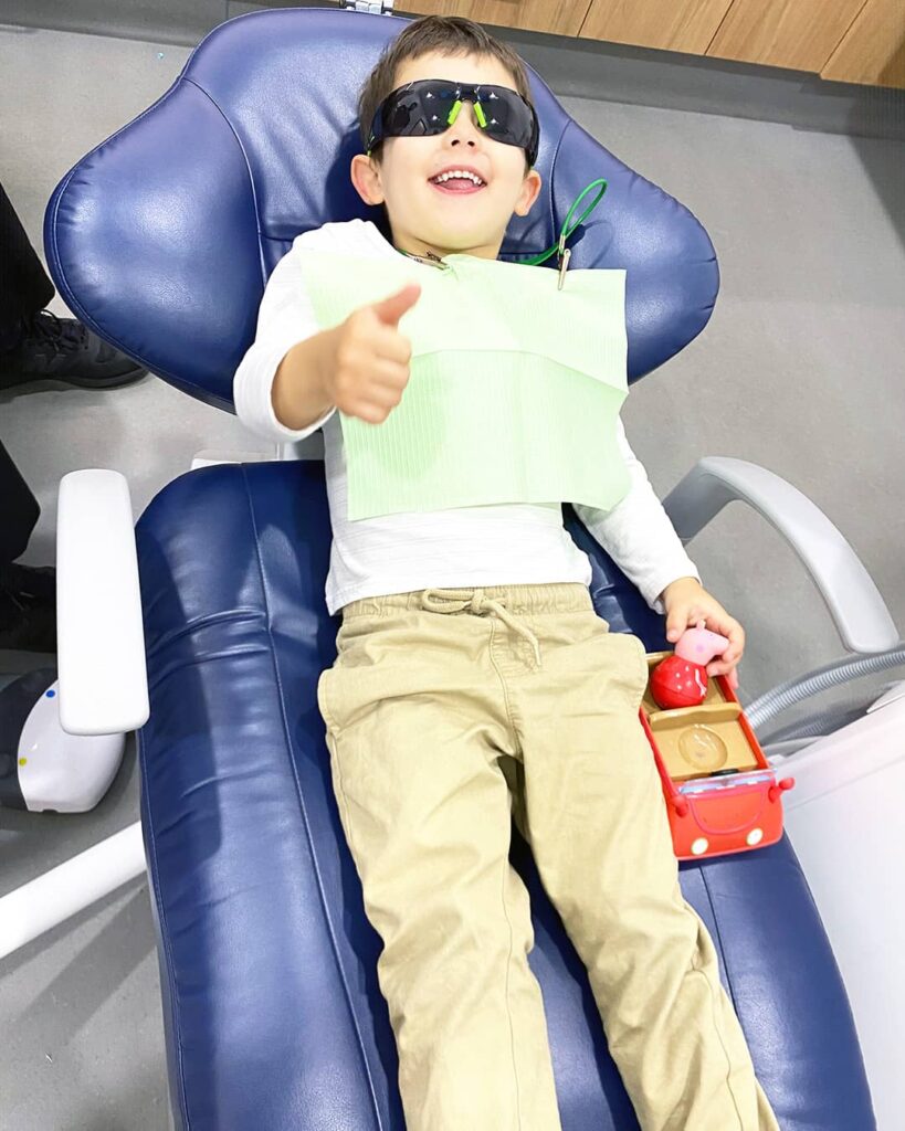 Happy boy patient wearing cool glasses showing OK sign in dental chair at Beyond Dental Care paediatric clinic