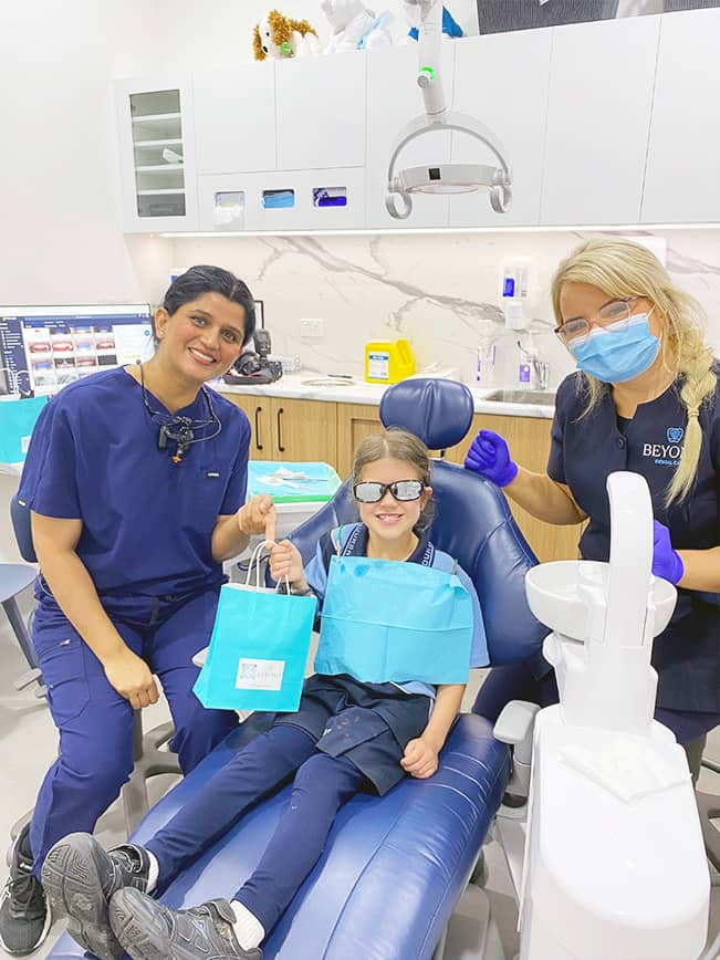 A young girl smiles after her dental check-up and cleaning at Beyond Dental Care, holding a gift bag with Dr. Puneeta Kumar and Practice Manager Emily Jackson.