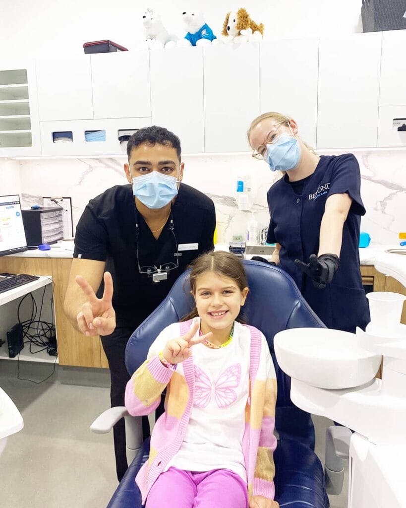 Our friendly paediatric dentist Dr Div and experienced dental assistant Paediatric dentist Dr Div and dental assistant smiling with happy girl patient showing OK sign at Beyond Dental Care clinic